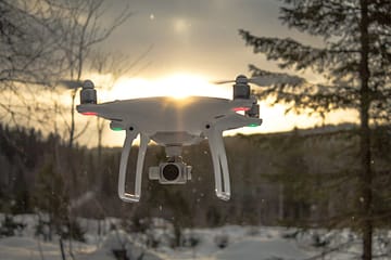 photo of white drone quadcopter flying near trees at noontime